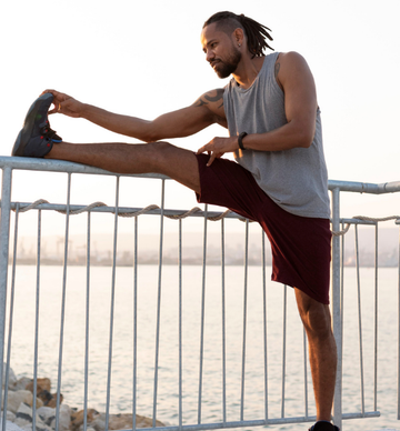 The image is a stock photo featuring an African American athlete performing a leg stretch on a pier or breakwater by the water, likely during sunrise or sunset. The man is wearing a gray tank top and maroon or burgundy athletic shorts.