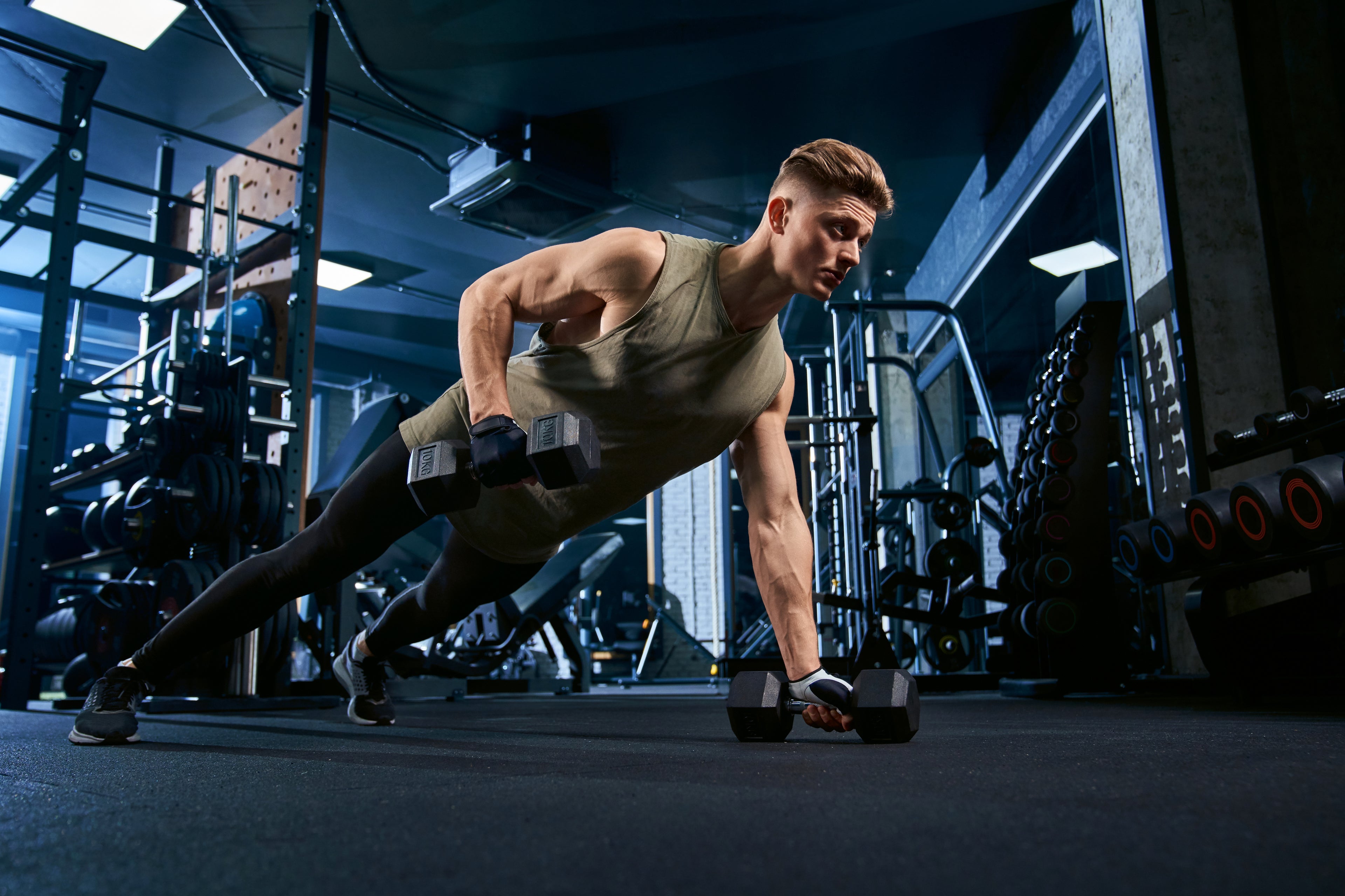 The image displays a man performing a dumbbell renegade row exercise in a gym setting. This is a compound exercise that works multiple muscles simultaneously. 
slider_item_YDit4y